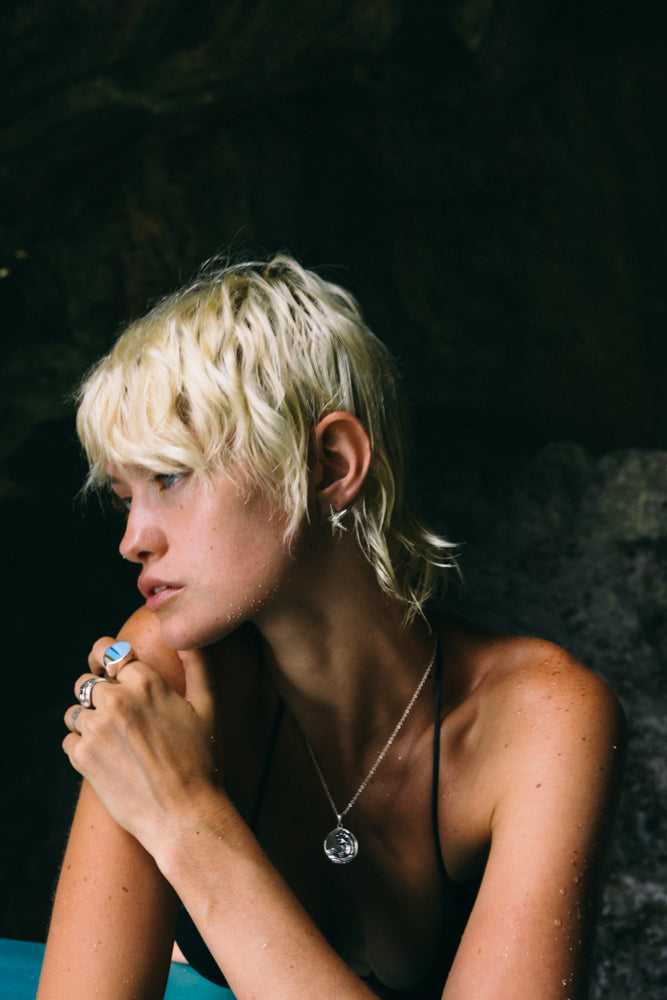 A person with short, tousled hair sits by a blue pool, wearing a black swimsuit and silver jewelry, surrounded by dark rock formations.