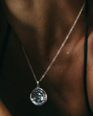 A person with short, tousled hair sits by a blue pool, wearing a black swimsuit and silver jewelry, surrounded by dark rock formations.