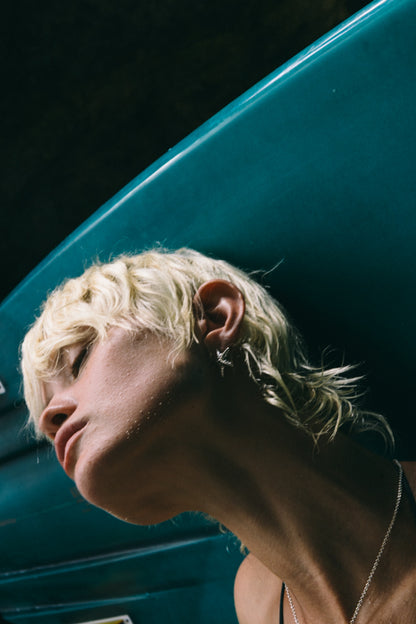 A close-up of a person with short, wavy blonde hair, facing sideways against a teal background, showcasing their neck and ear.