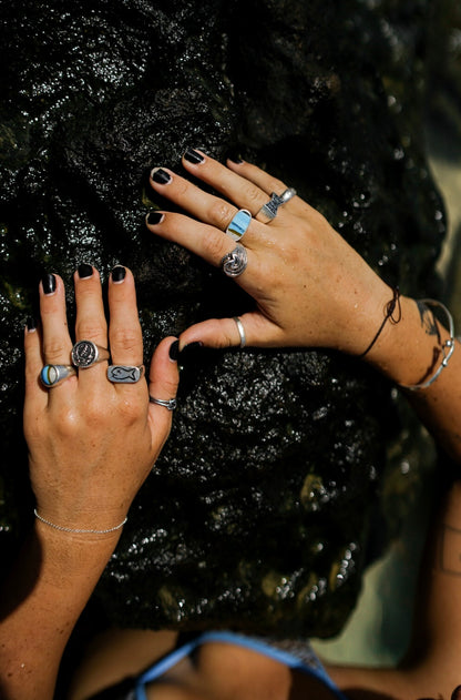 Close-up of two hands with black nails, adorned with various silver and gemstone rings, resting on textured dark rocks.