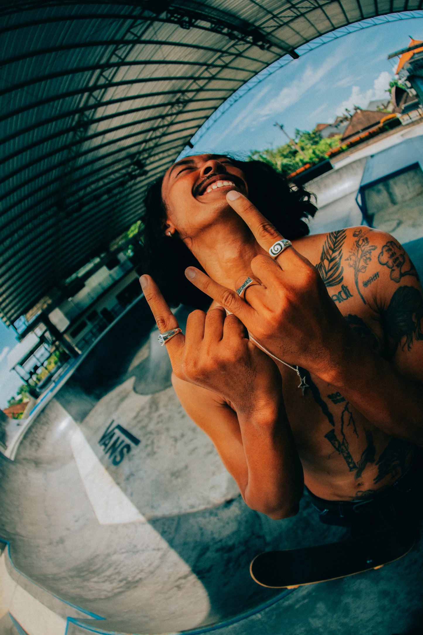 Model with Rock and Roll Hands at a skatepark smiling and looking into the distance. The model is showcasing our Handmade Jewelry Pieces. In the photo the model is wearing- the Spiral Black Sand Ring, Badfish Ring, Teach Peace Ring, and Rockstar Pendant on Silver Chain 