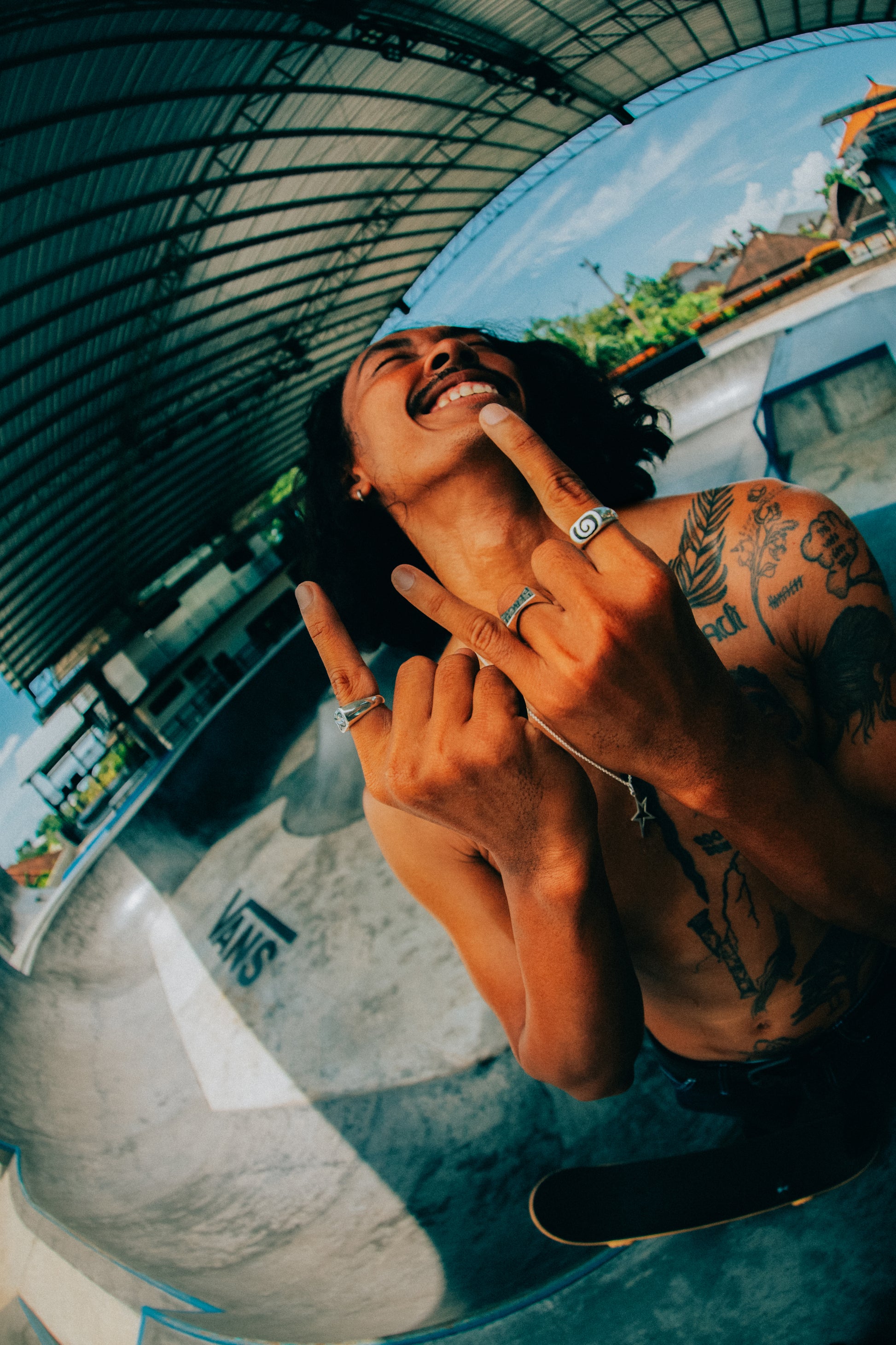 Model with Rock and Roll Hands at a skatepark smiling and looking into the distance. The model is showcasing our Handmade Jewelry Pieces. In the photo the model is wearing- the Spiral Black Sand Ring, Badfish Ring, Teach Peace Ring, and Rockstar Pendant on Silver Chain 