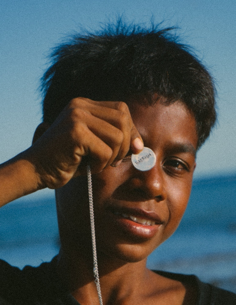 Little boy holding Tsunami Pendant Necklace in Silver, Highlighting the back of the Coin Pendant- it reads "Badfish" , the brands name. The front of the Pendant has a hand carved Wave Design on it. 